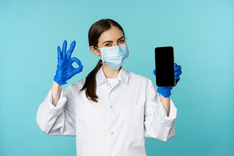 Portrait of doctor in medical face mask and gloves, showing mobile phone app, smartphone screen and okay sign, recommending online checkup website, standing over torquoise background.