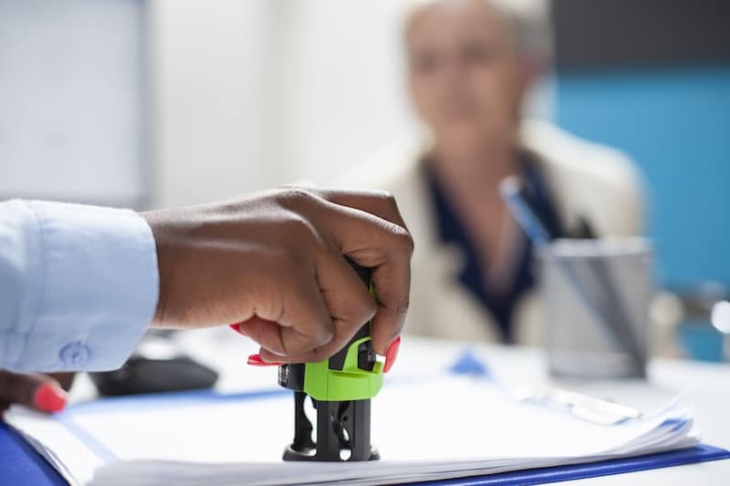Selective focus on hand of black healthcare specialist applying stamp on document as senior woman watches attentively. Elderly patient waits as african american doctor completes her medical documents.