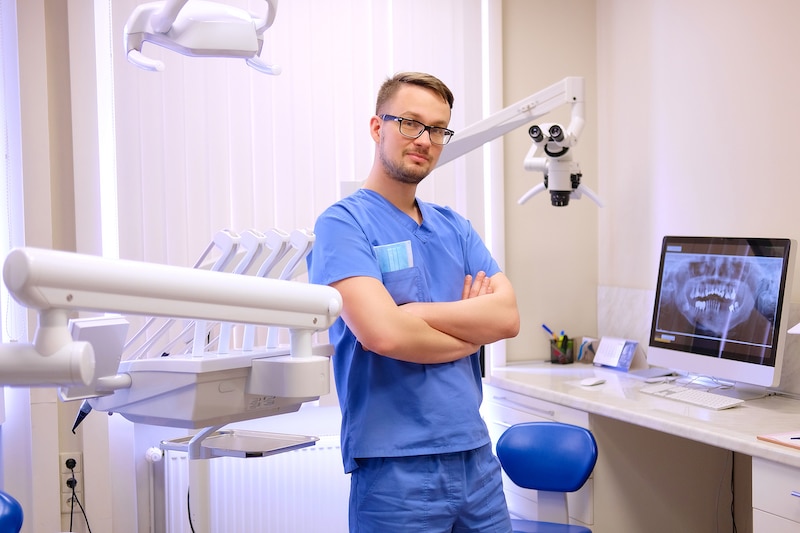 Portrait of a handsome dentist wearing a blue uniform, standing in a dentist clinic. Looking at camera.