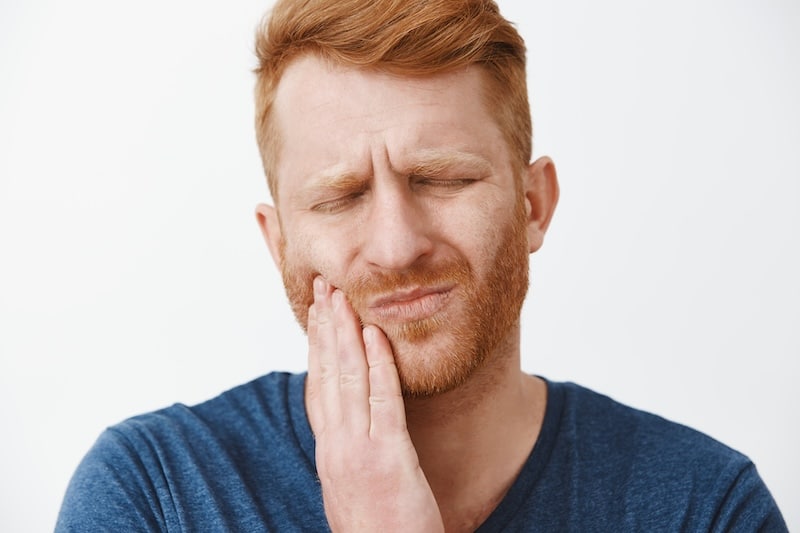 Close-up shot of redhead man with beard feeling pain in teeth, frowning and making suffering expression with closed eyes, touching cheek, having need to call dentist for curing decay or rotten tooth. Health and dental problems concept